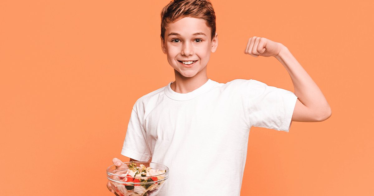 young boy wearing white shirt infront of orange background showing his muscle with bowl of nutrients