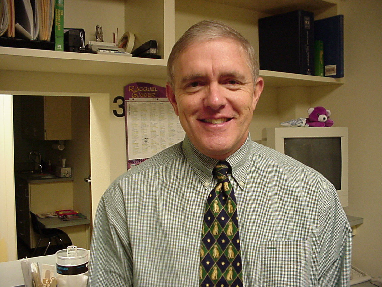 A man with short gray hair, wearing a green checkered shirt and a patterned tie, stands smiling in an office with shelves, papers, and a computer monitor in the background.