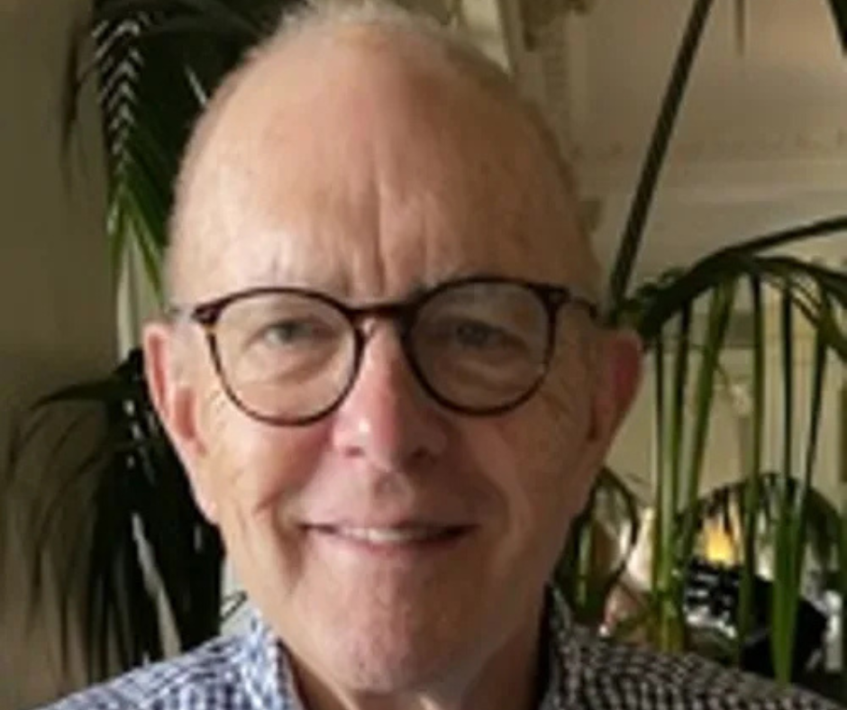 An older man with glasses and a slight smile is indoors, standing in front of green plants. He wears a blue and white checkered shirt.