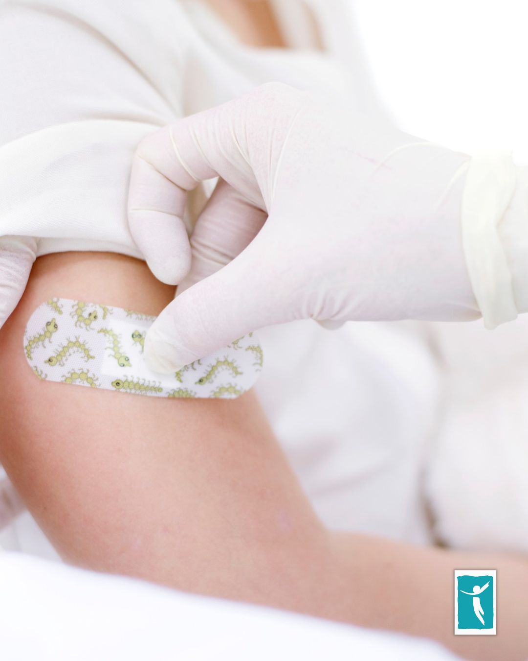A gloved hand places a bandage with cartoon designs on a person's upper arm, likely after receiving the covid-19 vaccine; the person is wearing a white shirt.