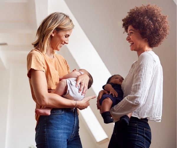 Two women are standing and smiling at each other while each holds a baby in their arms, in a bright indoor setting. The atmosphere appears warm and friendly.