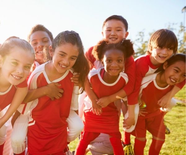 A group of smiling children in red soccer uniforms stand close together outdoors on a sunny day, some giving piggyback rides, appearing happy and playful.