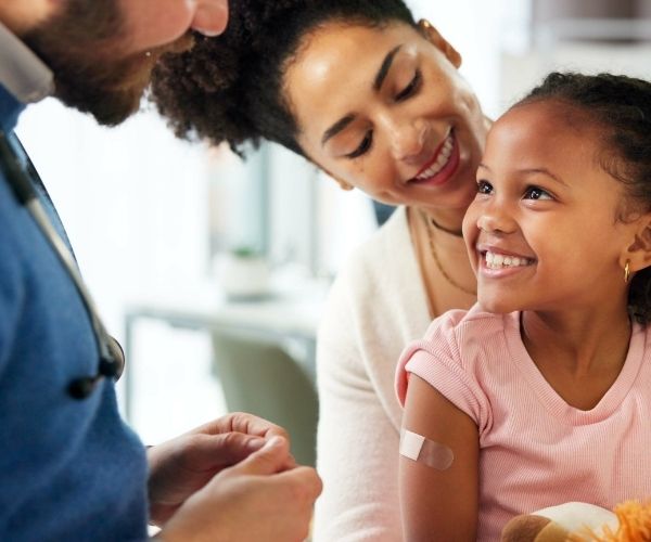 A smiling young girl with a bandage on her upper arm sits on her mother's lap, looking happily at a healthcare professional after receiving a vaccination. The mother smiles warmly at her daughter.