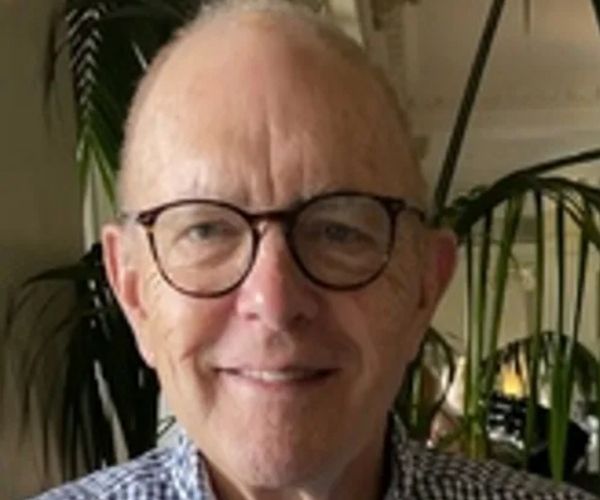A smiling older man with glasses and a checkered shirt sits indoors with green plants and a white ceiling in the background.