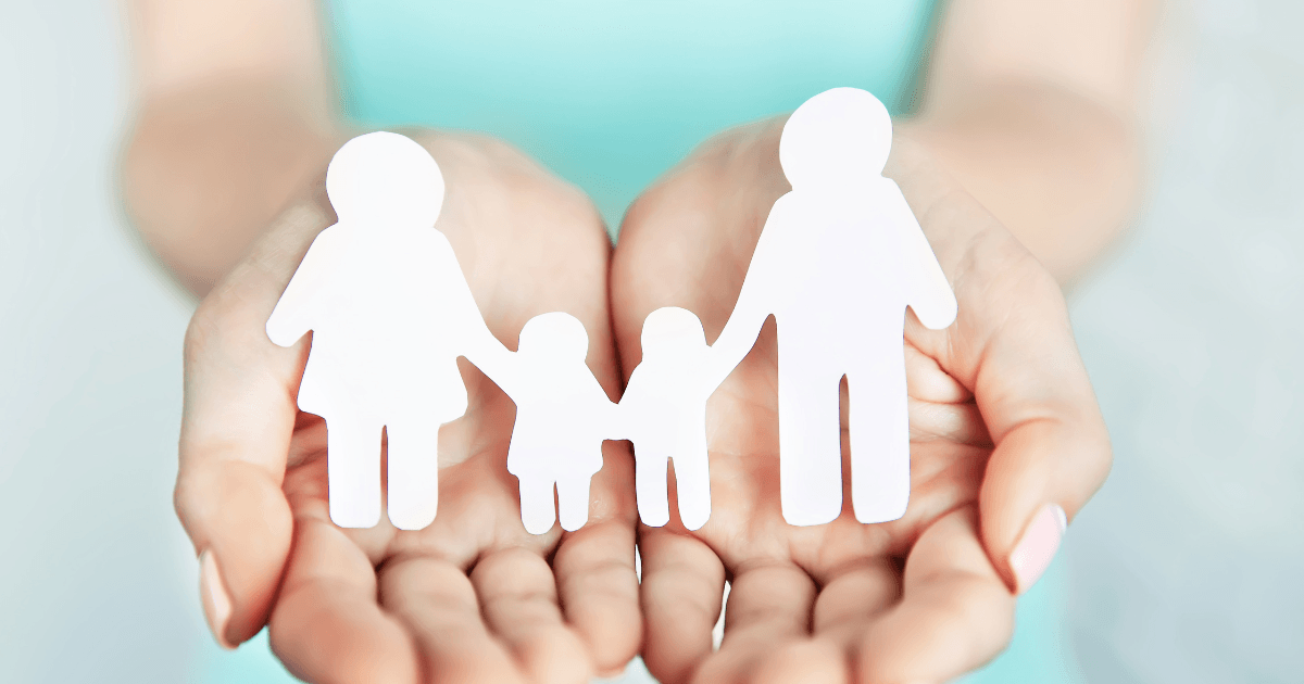 A person holds paper cutouts of a family—two adults and two children—on their outstretched palms, symbolizing care and protection of the family unit.
