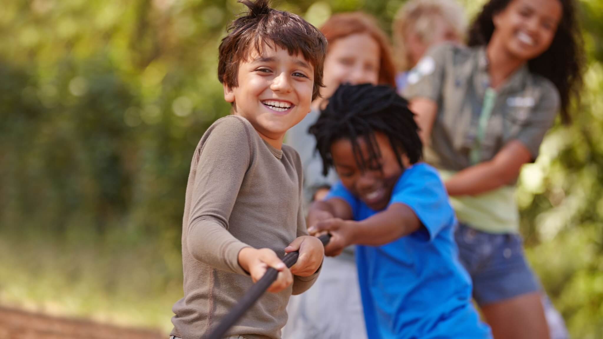 Kids Playing at Camp In Colorado Children smiling and playing tug-of-war outdoors, meeting summer camp physical requirements as they pull on a rope together with greenery in the background. The atmosphere is joyful and energetic.